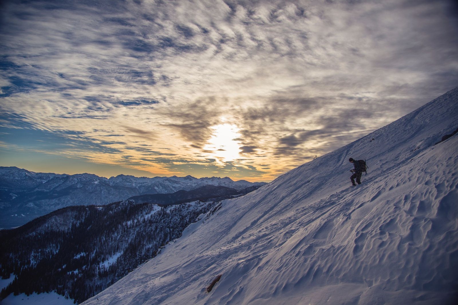 A climber crossing a steep snow slope at dawn above a valley of forested ridges near Radovljica