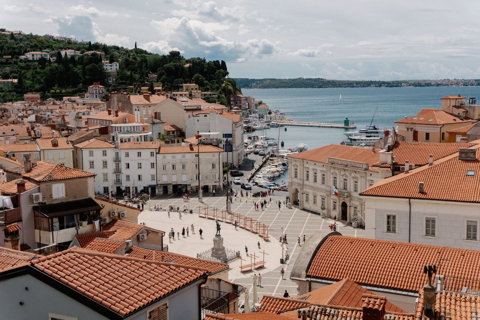Tartini Square in Piran from above, an oval of pale stone ringed by red-roofed houses, with the harbour and the sea beyond
