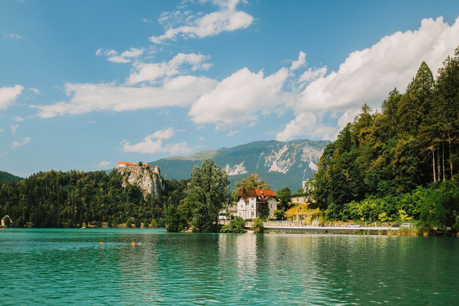 Lake Bled in summer, green water in the foreground, a villa among trees on the shore and the castle on its cliff behind