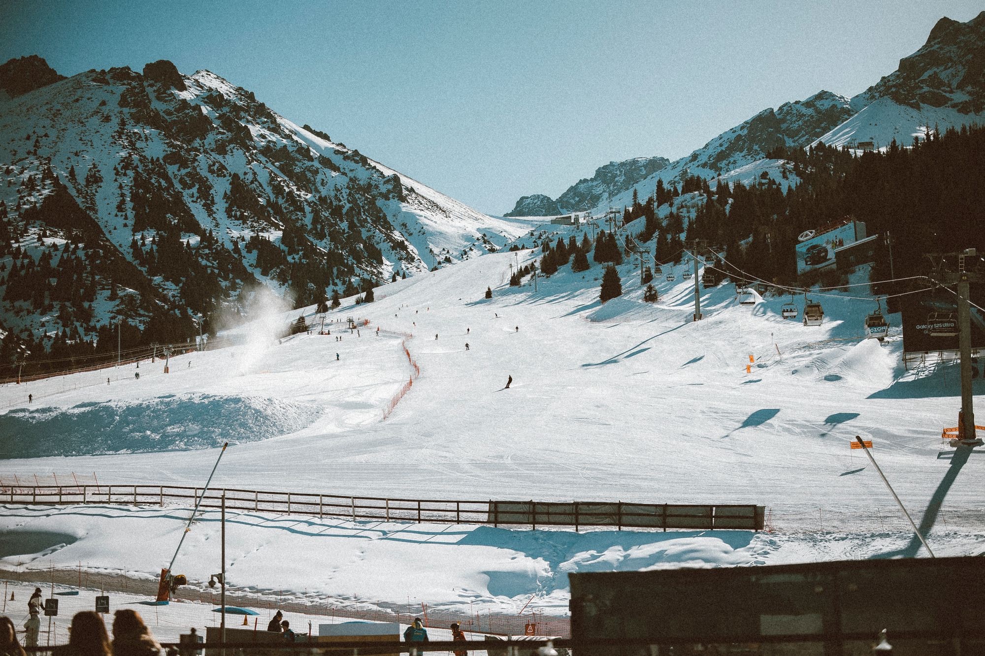Skiers on the main slope above the Shymbulak base, with chairlifts on the right