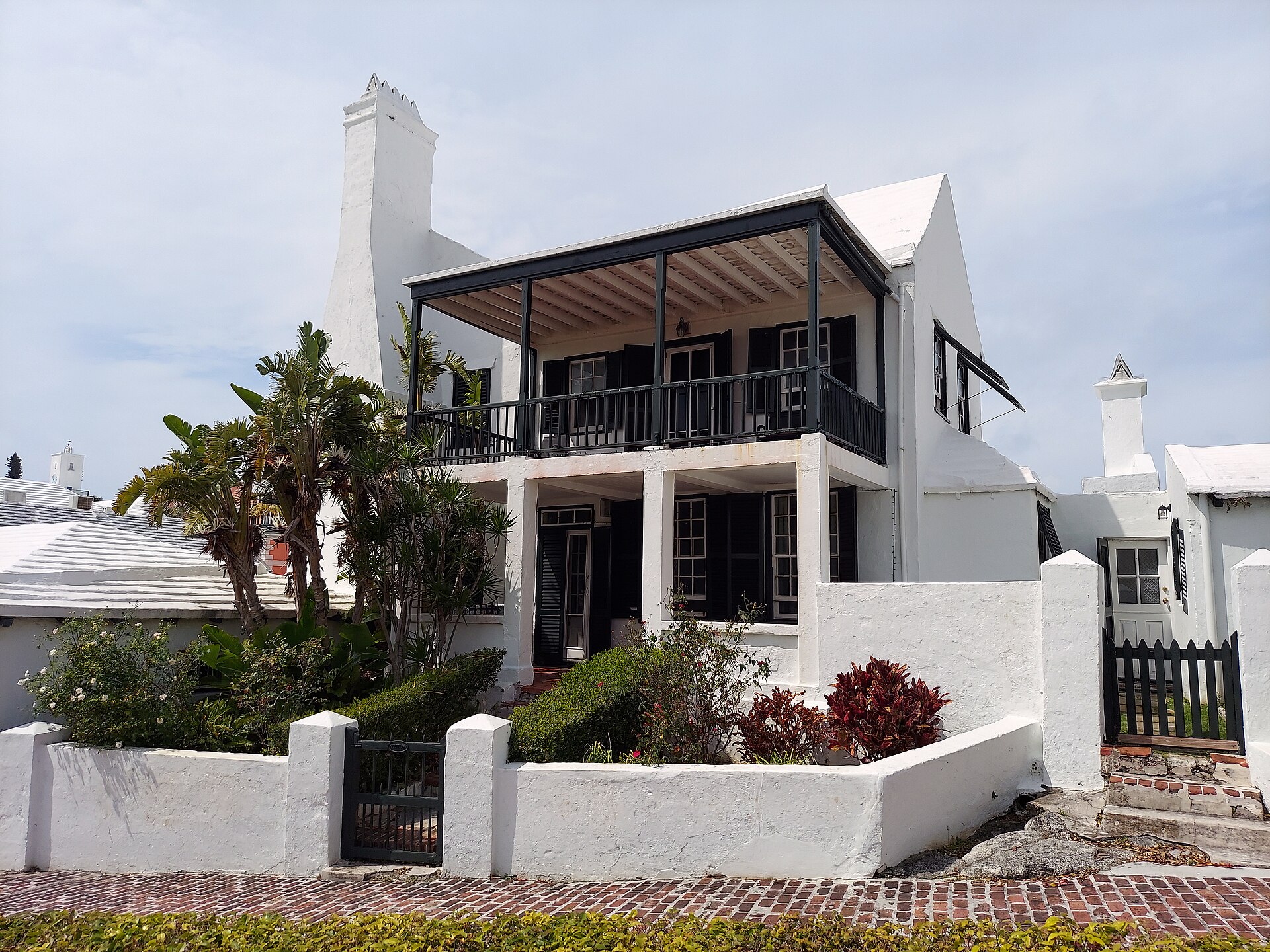 A white Bermudian house with a deep veranda, dark shutters and a stepped white limestone roof, behind a low white wall with tropical planting