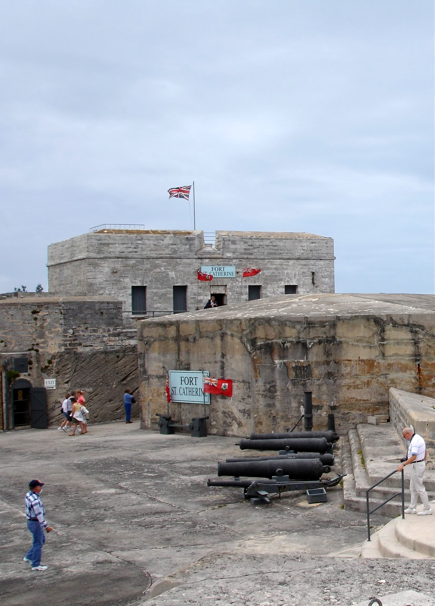 A pale stone fort with a flagpole and a row of black cannon on a paved terrace, a few visitors walking below the ramparts