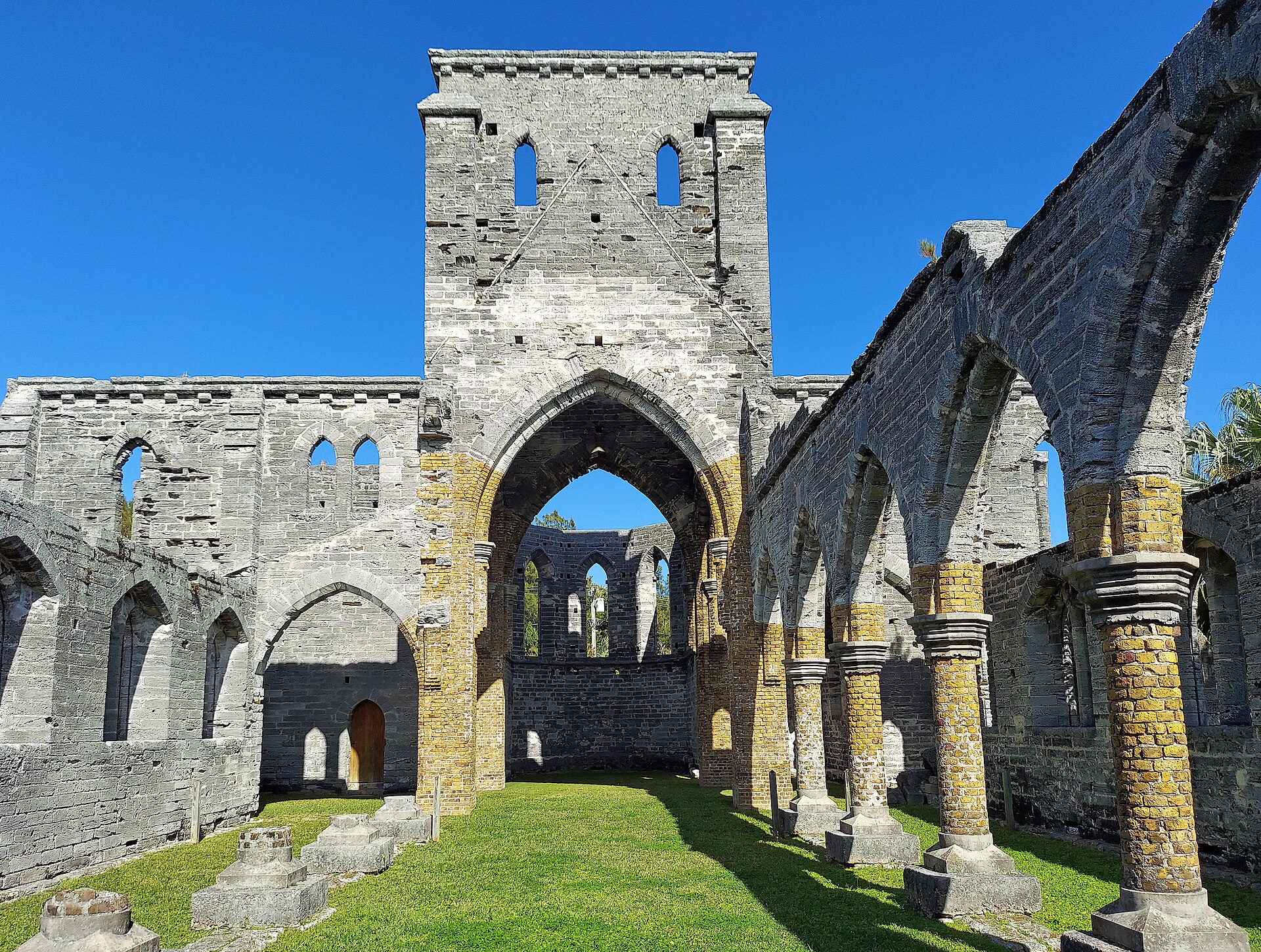 The roofless interior of a Gothic limestone church, its arcade of pointed arches and empty window openings standing open to a blue sky, grass growing on the floor