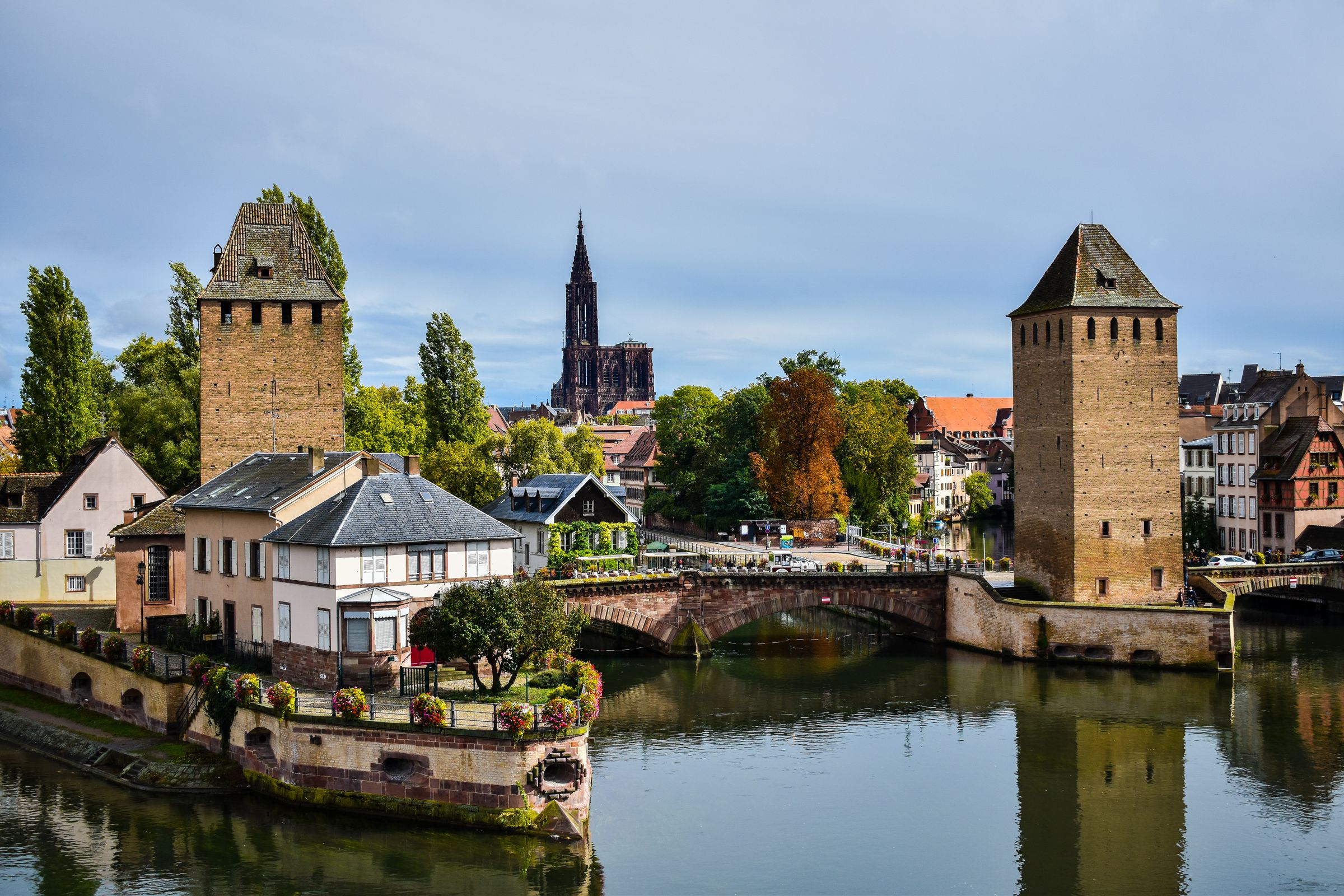 The two square stone towers of the Ponts Couverts over the still Ill, a stone arched bridge between them, with the dark red sandstone spire of the cathedral rising behind the trees and roofs and flower boxes along a waterside wall on the left