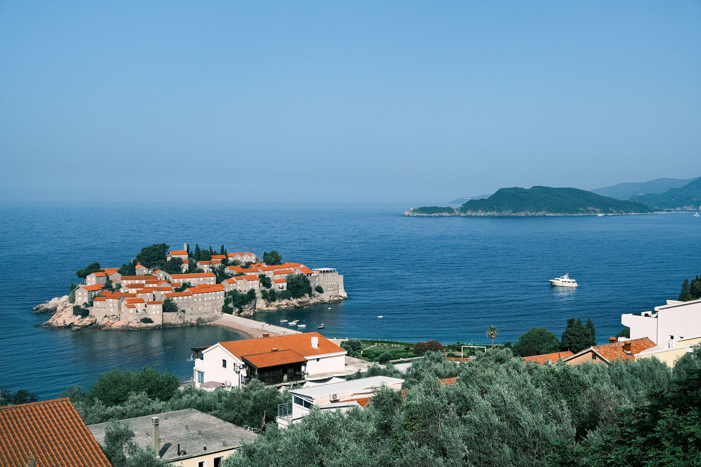 Sveti Stefan's walled island village on its causeway in a calm blue sea, seen from the wooded hillside above
