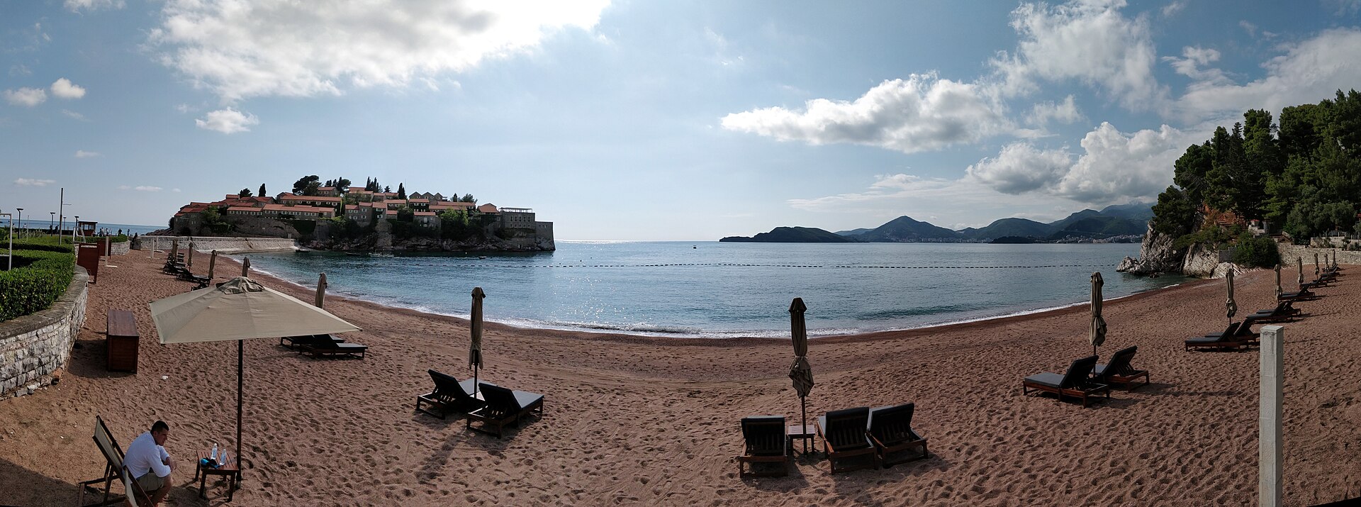 A wide bay north of Sveti Stefan, scattered sun loungers and furled umbrellas on the pebble beach, the island at the far left