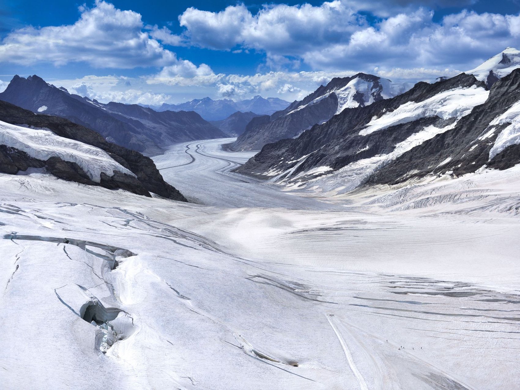 A wide white glacier filling a valley between dark rock ridges under a blue sky, with crevasse lines curving down its length