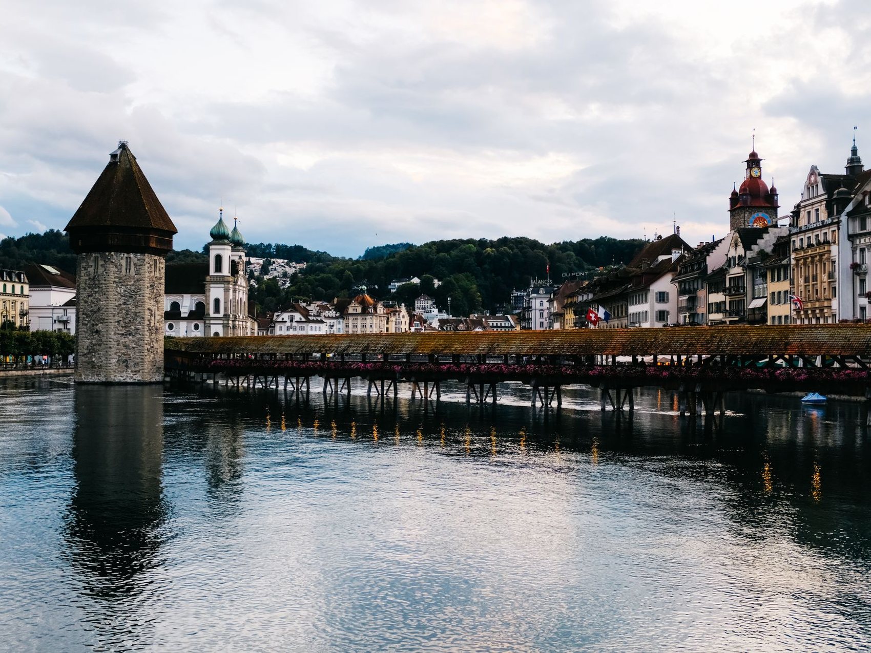 A long covered wooden bridge crossing a river towards a stone water tower, with the spires and painted facades of a lakeside town behind it