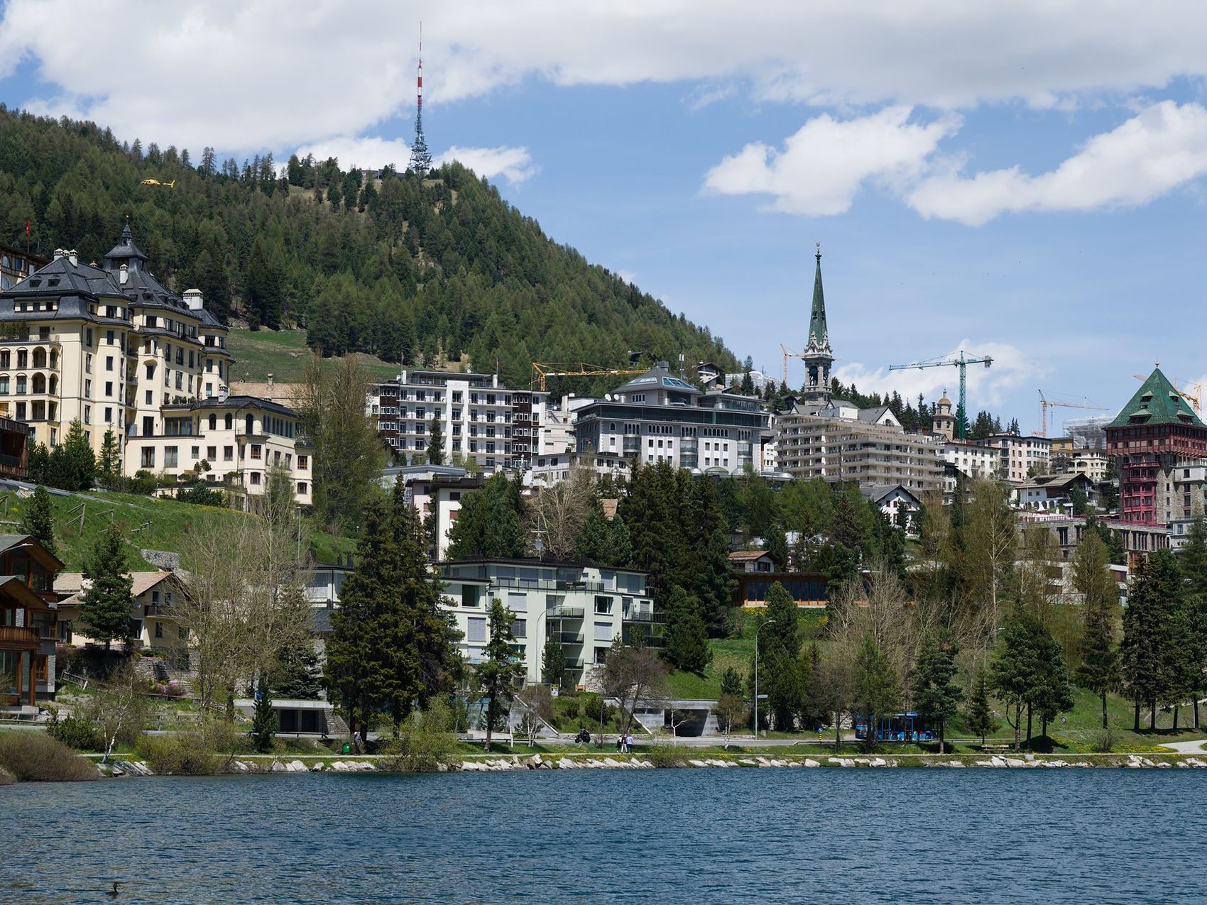 A hillside town of large pale hotels and a church spire above a lake, with wooded slopes and a communications mast on the ridge behind