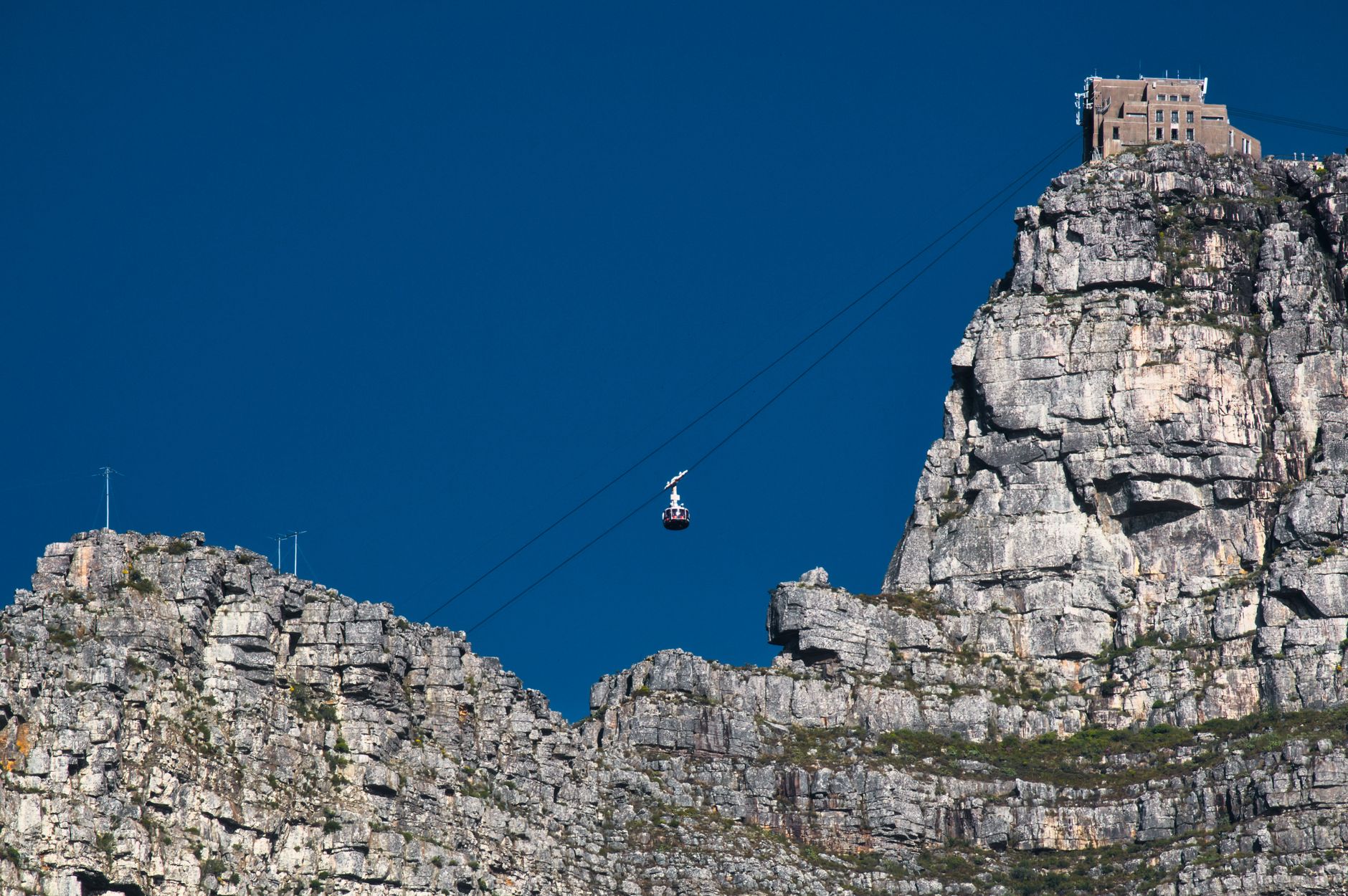 A single round cable car hanging mid-span below the upper cableway station, which sits on the cliff edge above layered sandstone