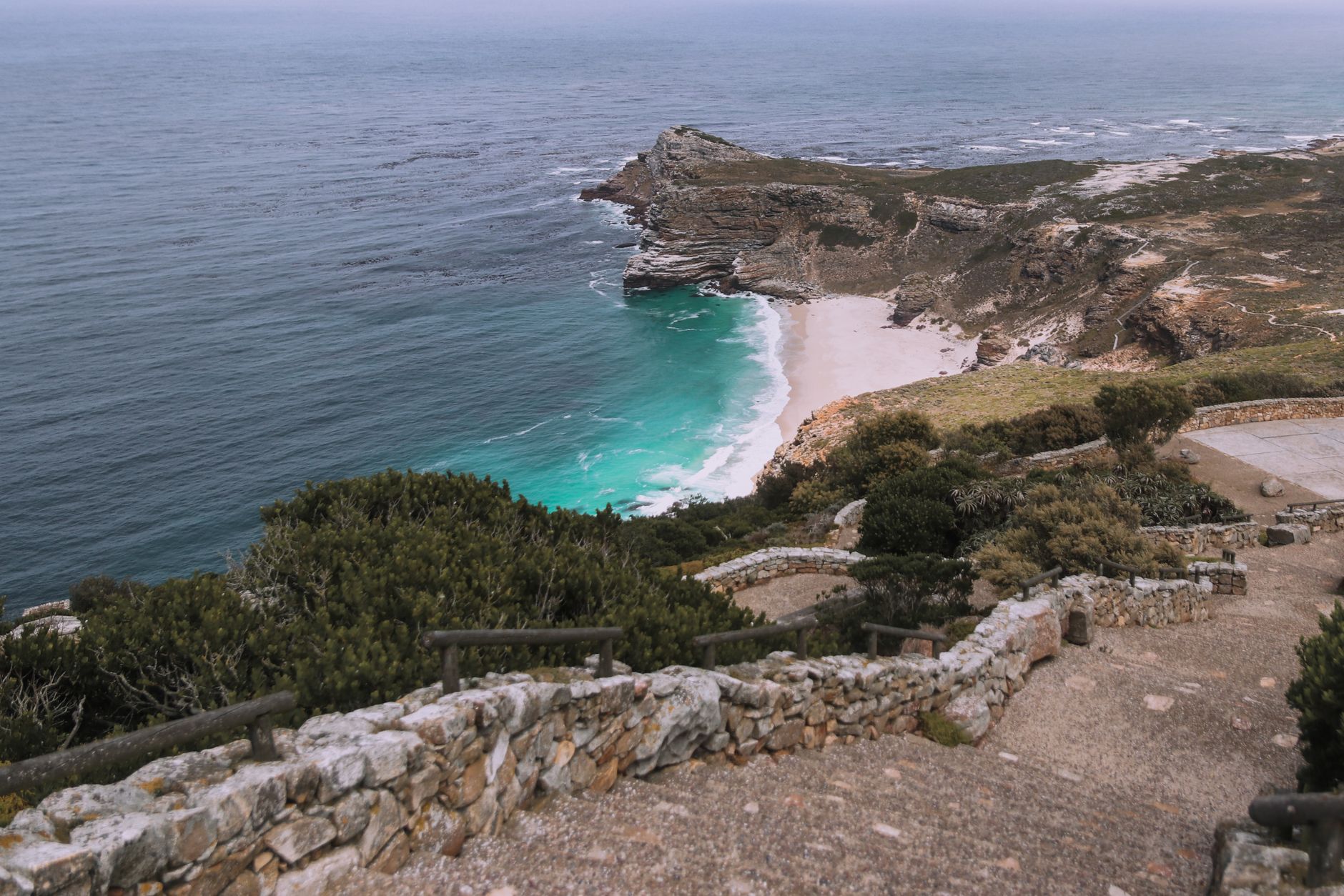 A stone-walled viewpoint path with timber railings in the foreground, looking down on a rocky headland, a crescent of white sand and turquoise water