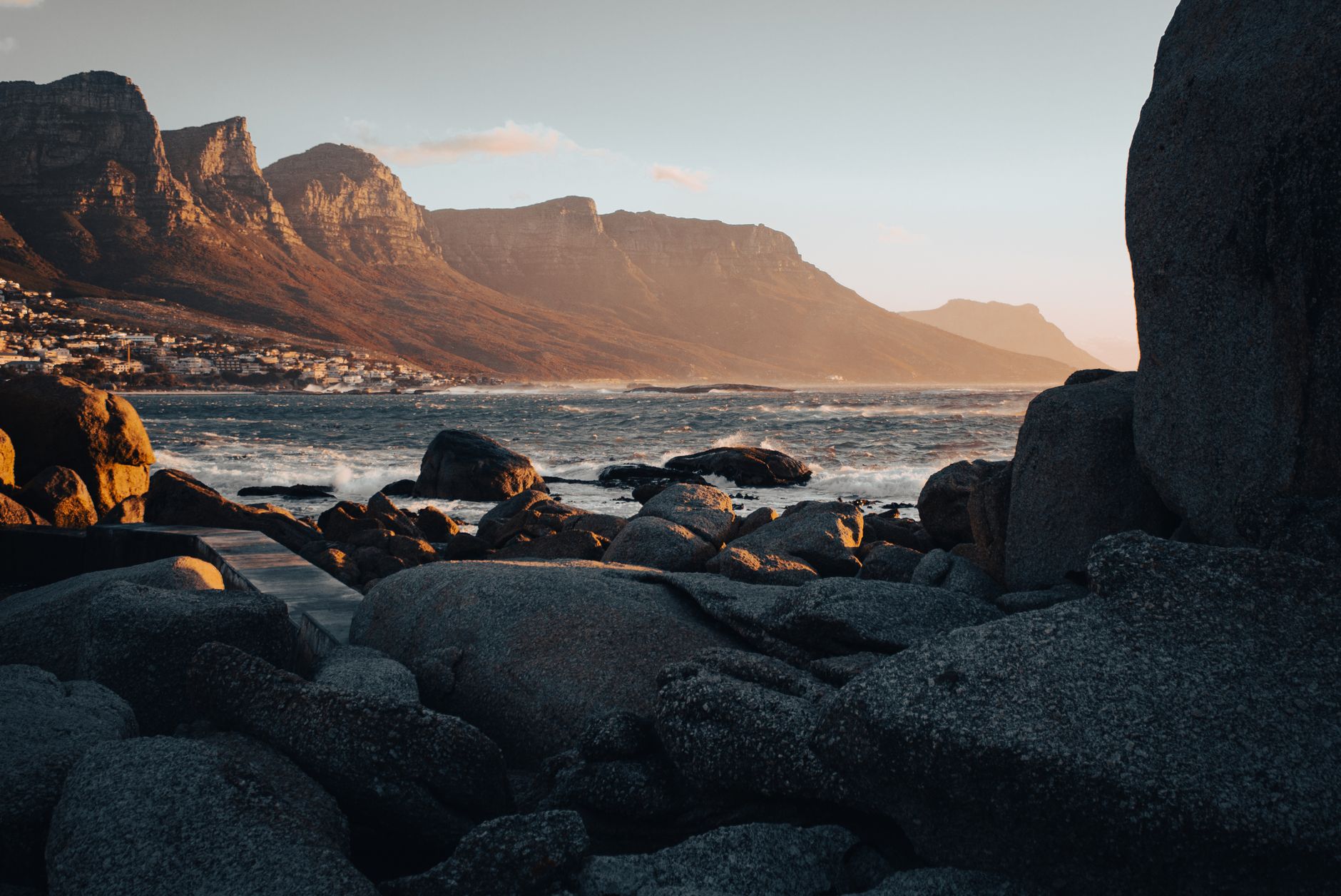 A line of buttressed sandstone cliffs in low golden light above a coastal suburb, with large rounded granite boulders on the shore in the foreground