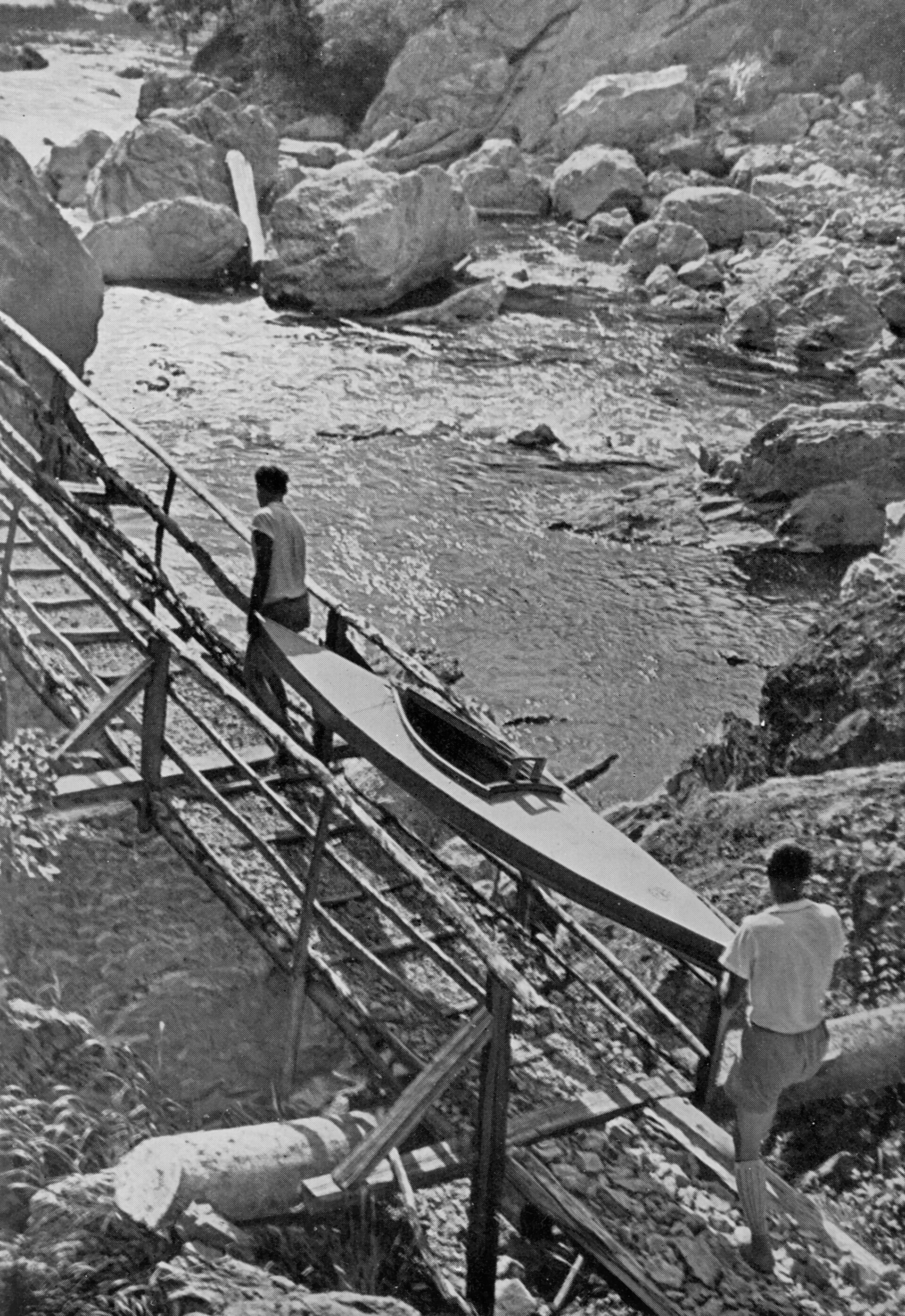 A 1932 photograph of two men carrying a folding kayak along a timber walkway built out over the boulders of the Tara