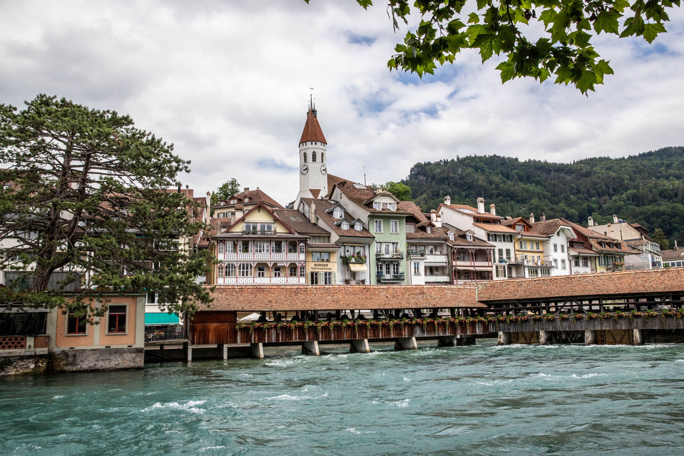 Thun's covered wooden lock bridge over the fast green Aare, with old town houses and the clock tower of the town church behind
