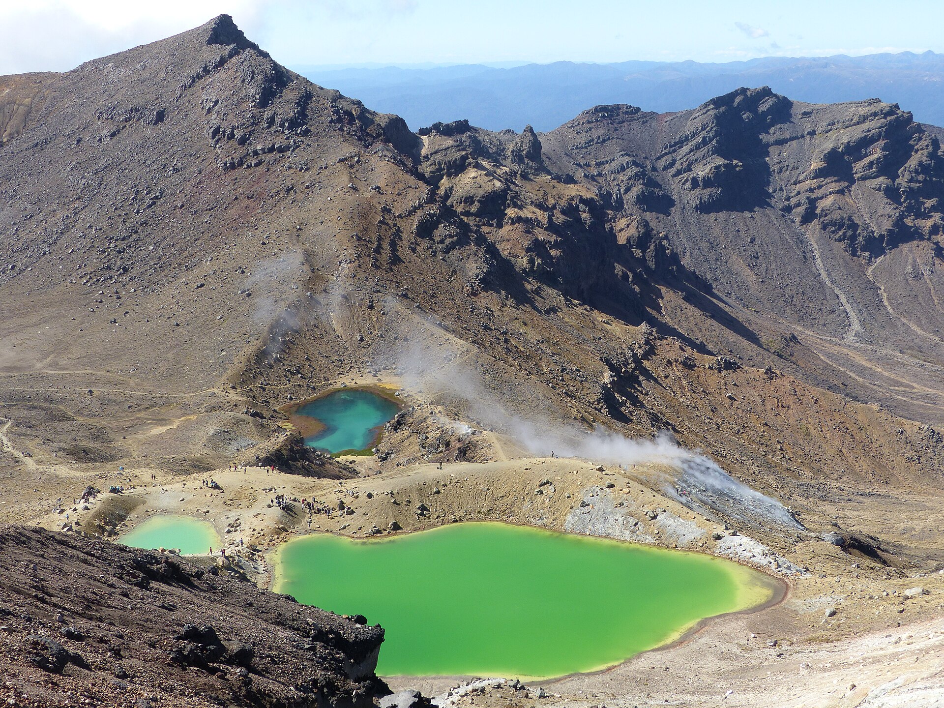 Three vivid green crater lakes below a grey volcanic ridge with steam rising