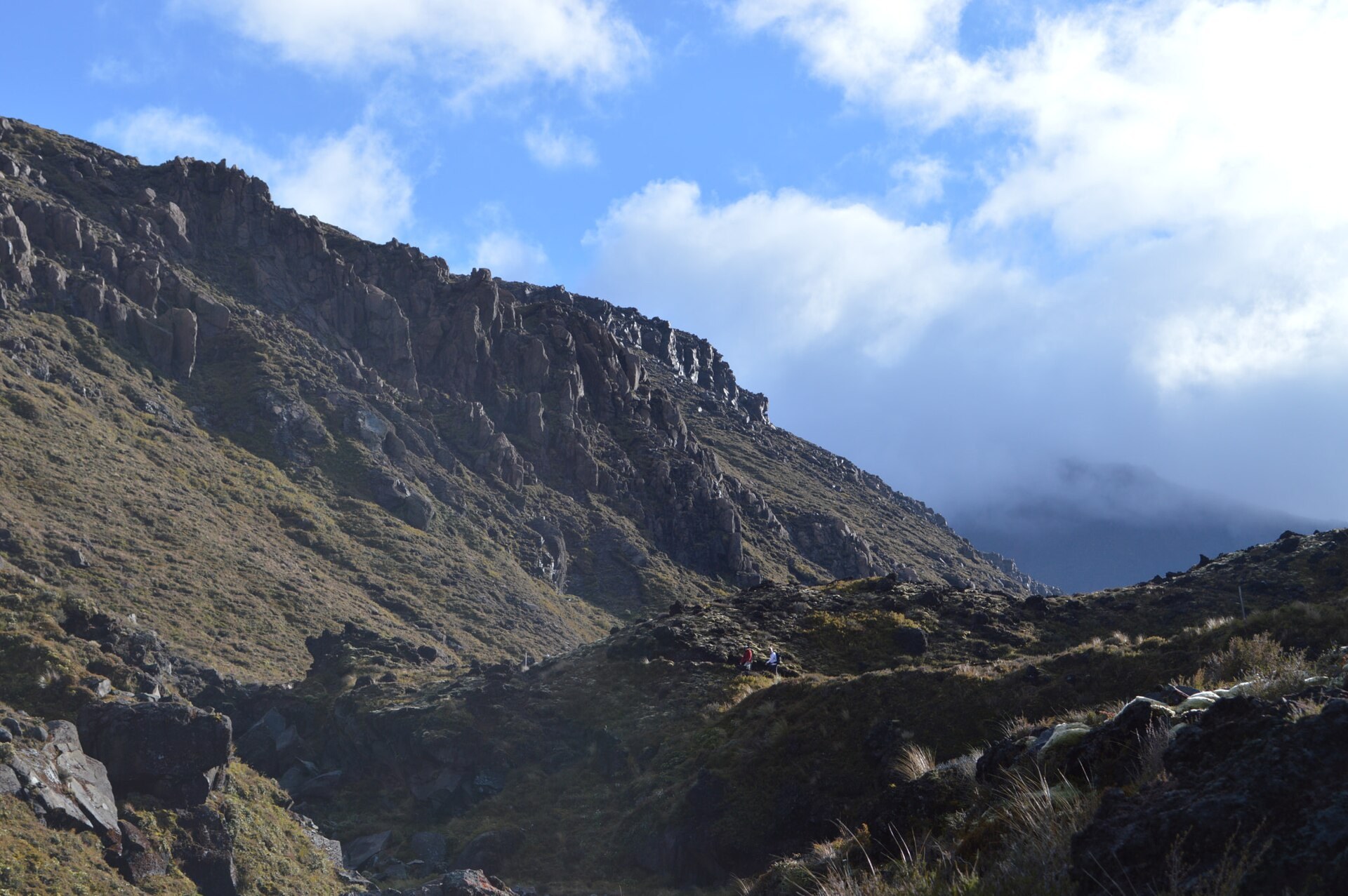 Two walkers on the track through the Mangatepopo valley below a dark rock ridge