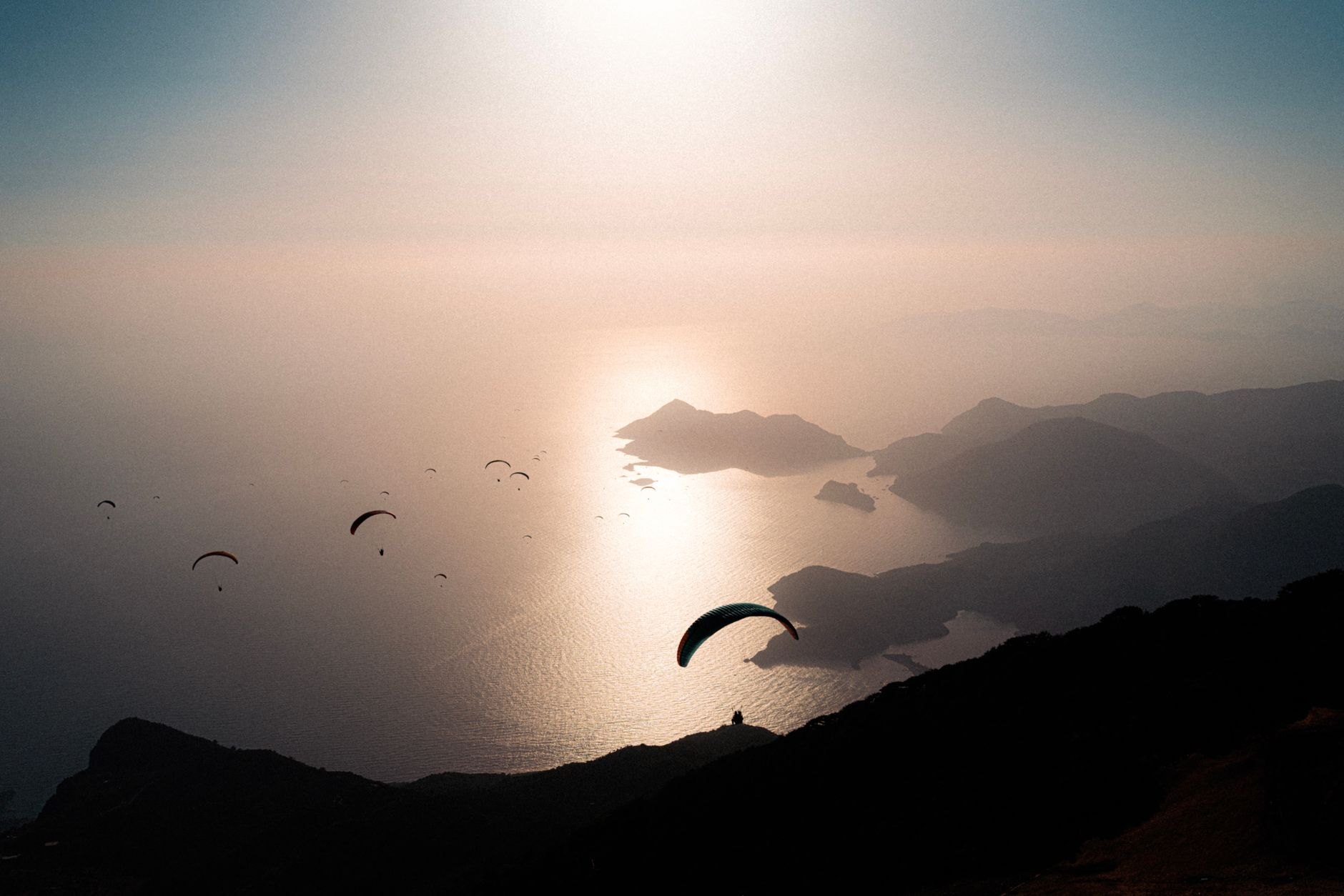 A dozen paragliders in silhouette stacked above a hazy sea at dusk, with headlands and the sun low over the water