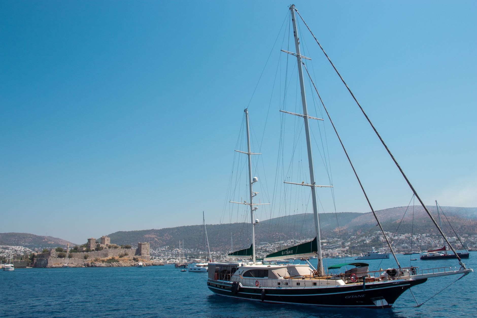 A wooden gulet under sail on flat blue water off Bodrum, with the towers of the crusader castle on the headland behind it