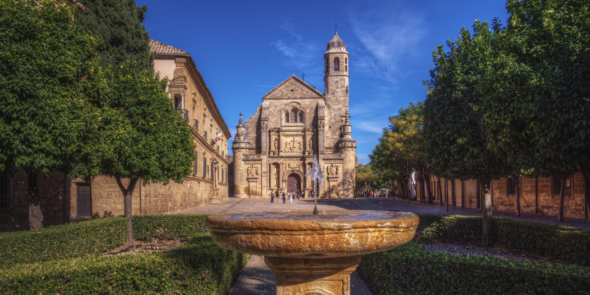 The carved front and bell tower of the Sacra Capilla del Salvador at the end of Plaza Vázquez de Molina, with orange trees and a stone fountain in the foreground