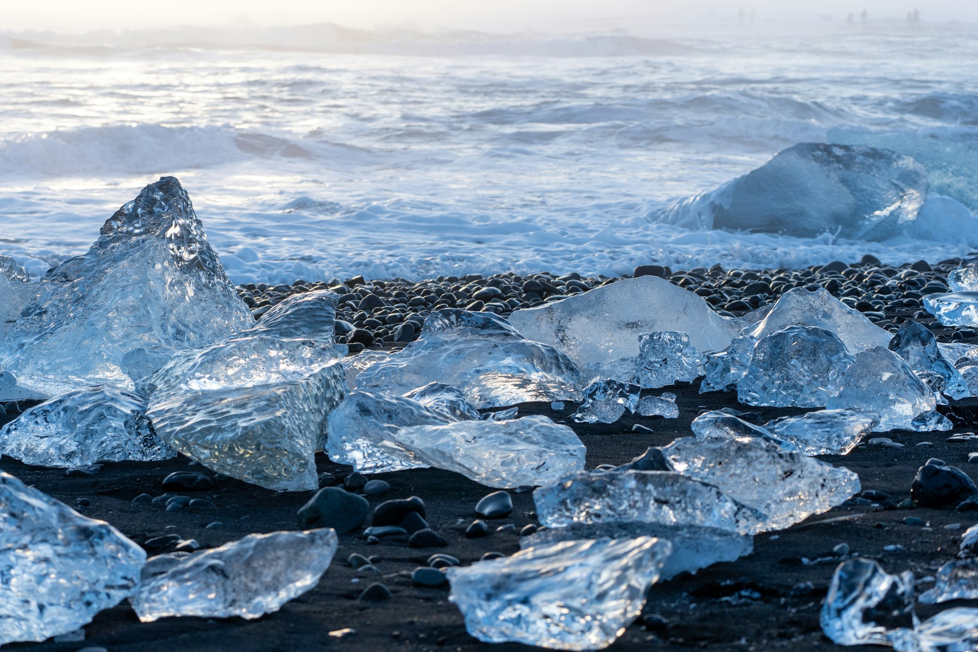 Clear blue blocks of ice strewn across black sand with surf breaking close behind in low light