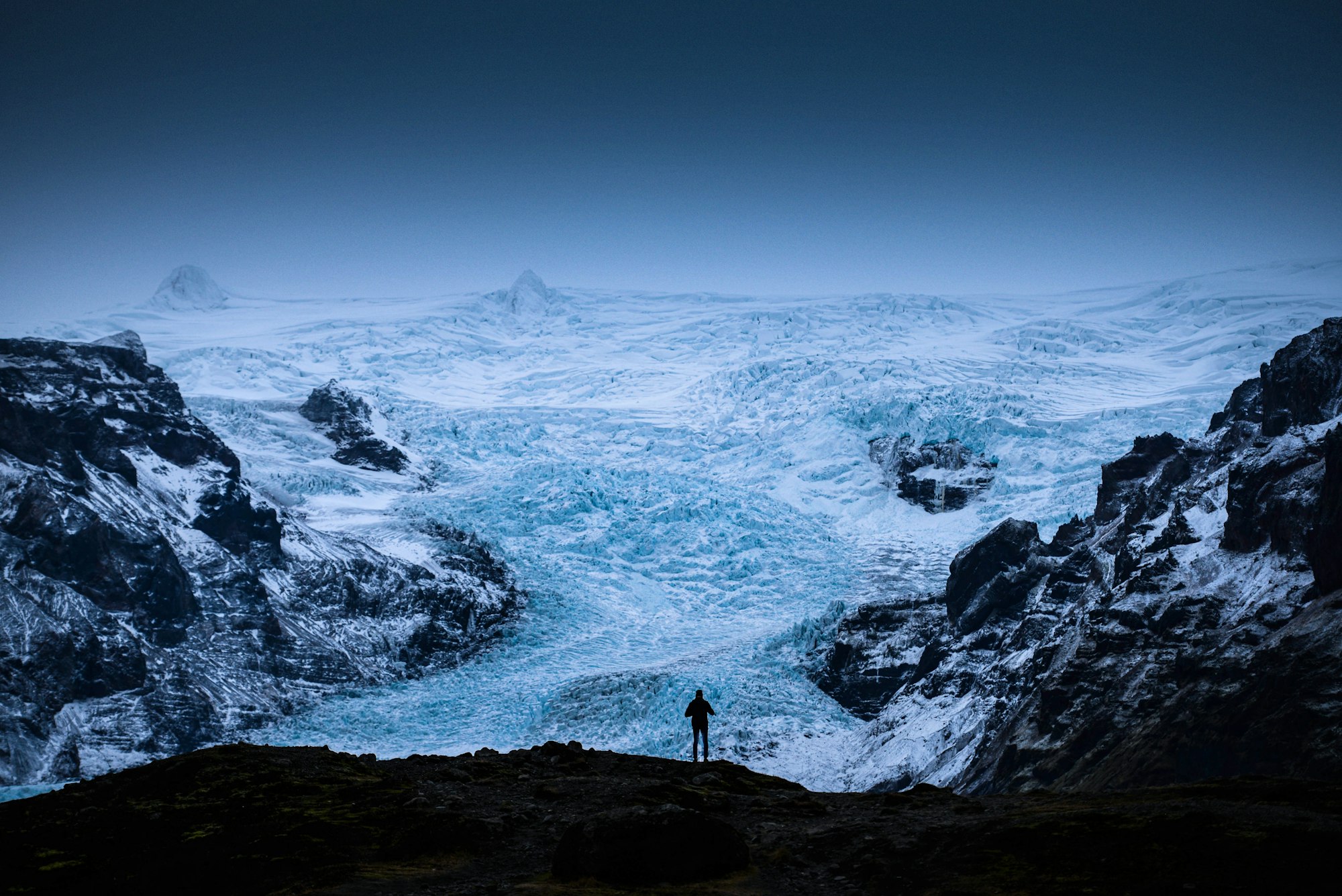 A single figure standing on dark rock looking out over a vast crevassed glacier tongue between rock walls in flat light