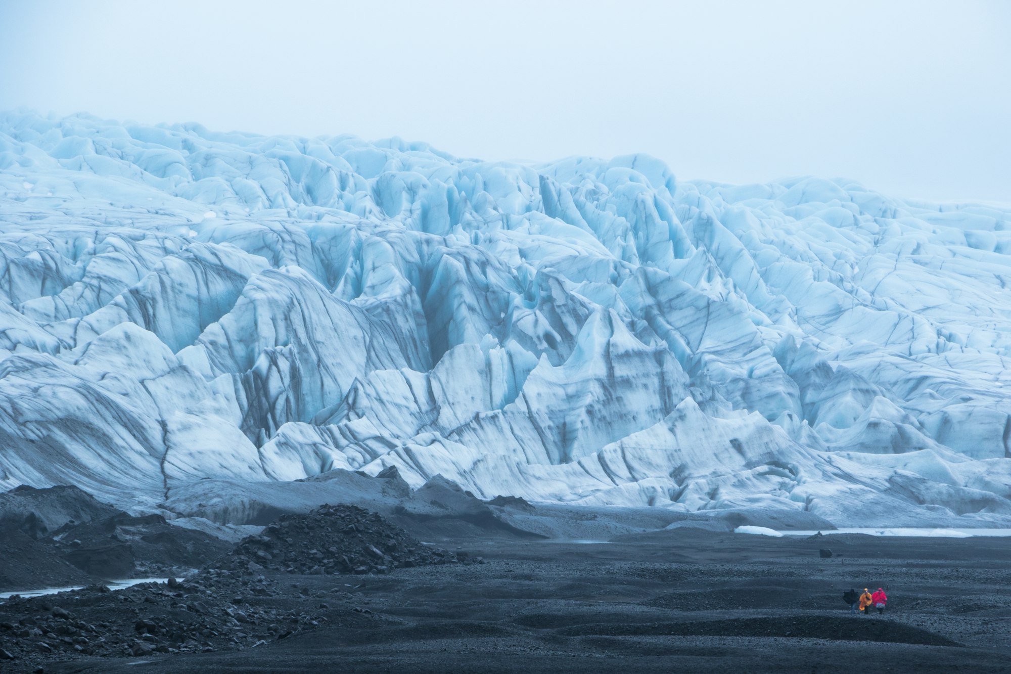 A wide crevassed glacier front rising from a dark moraine plain with two tiny walkers, one in orange and one in red, at the right edge
