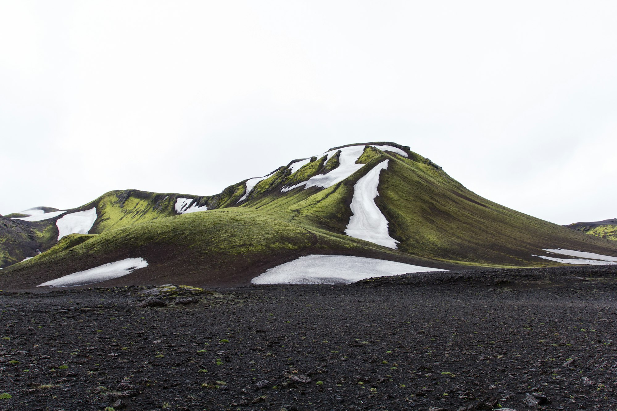A moss green volcanic hill with snow filled gullies rising from a flat black tephra plain under white overcast