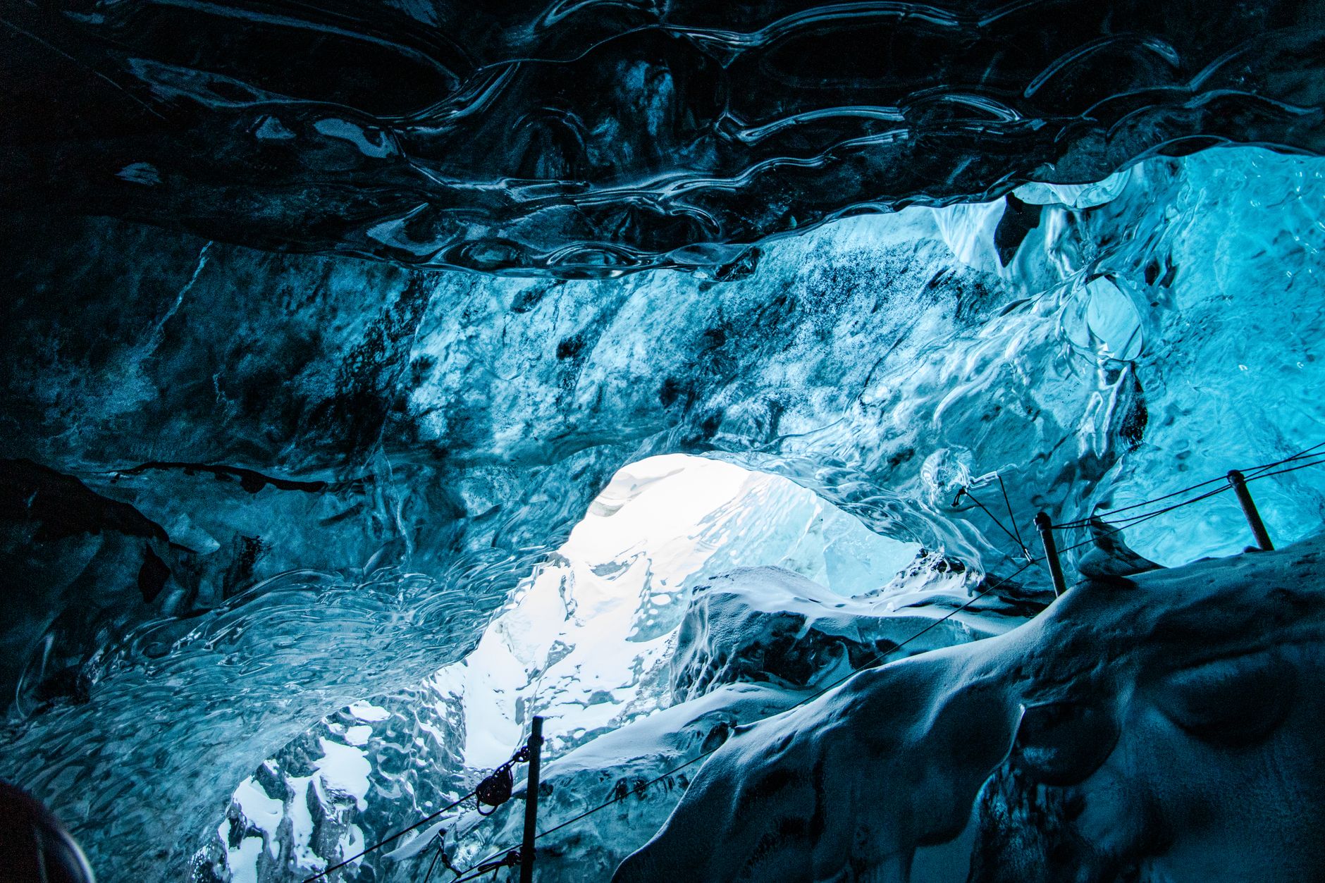 The interior of a natural ice cave with dark ash-laden ice in the roof, blue walls and a fixed safety rope on stanchions