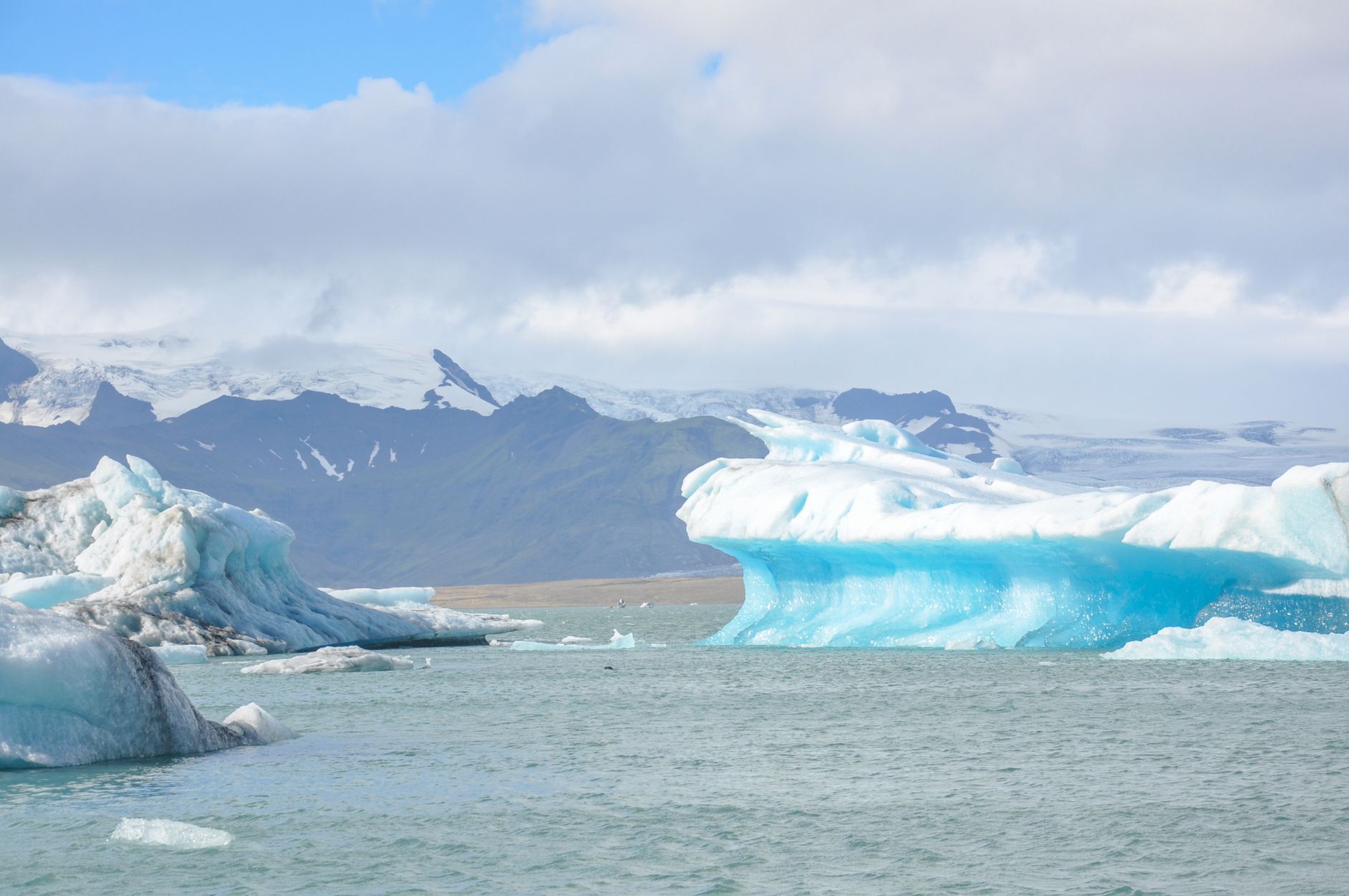 Blue and white icebergs on the wind ruffled water of Jokulsarlon glacier lagoon with snow covered mountains and the edge of the ice cap behind under broken cloud
