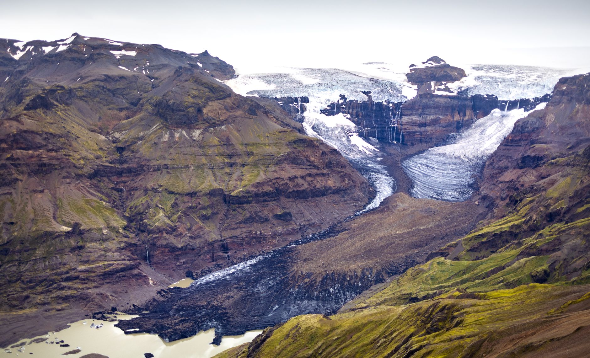 Aerial view of a glacial valley at Skaftafell with an icefall spilling over a cliff band into a debris covered tongue above a silt brown lake