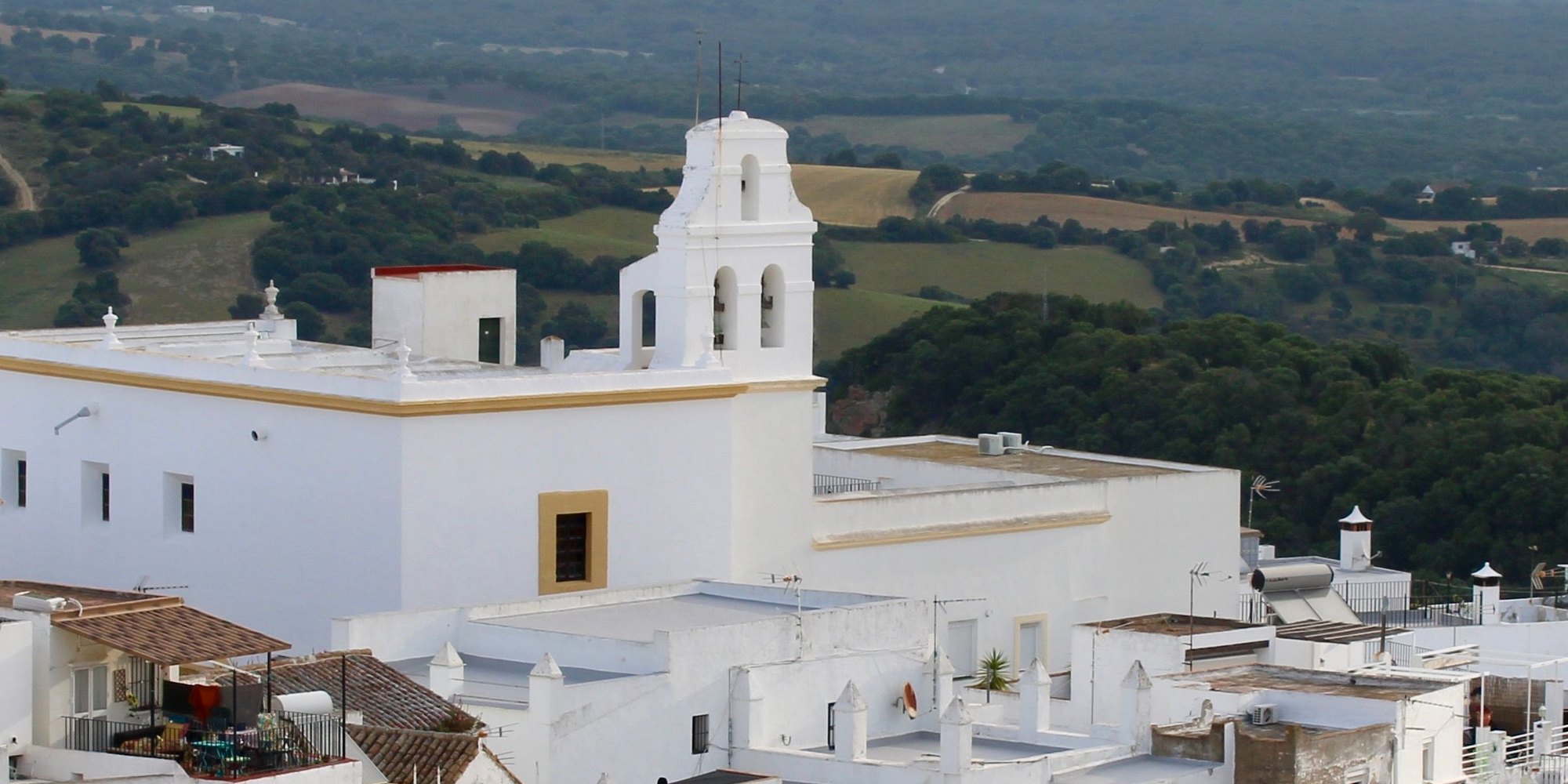 White rooftops and a bell gable in Vejer de la Frontera, with green farmland and woods stretching away below the hill