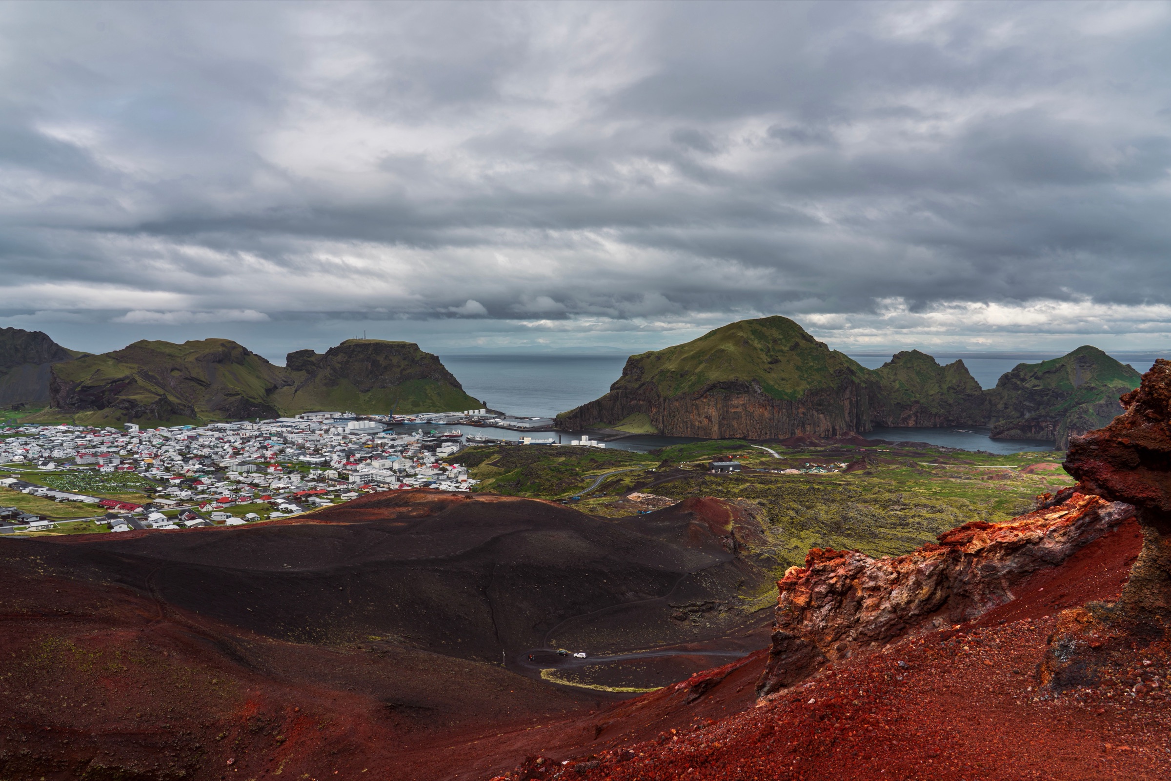 The town and harbour of Heimaey seen from the red scoria slopes of Eldfell, with the green cliffs of Heimaklettur across the water under a grey sky
