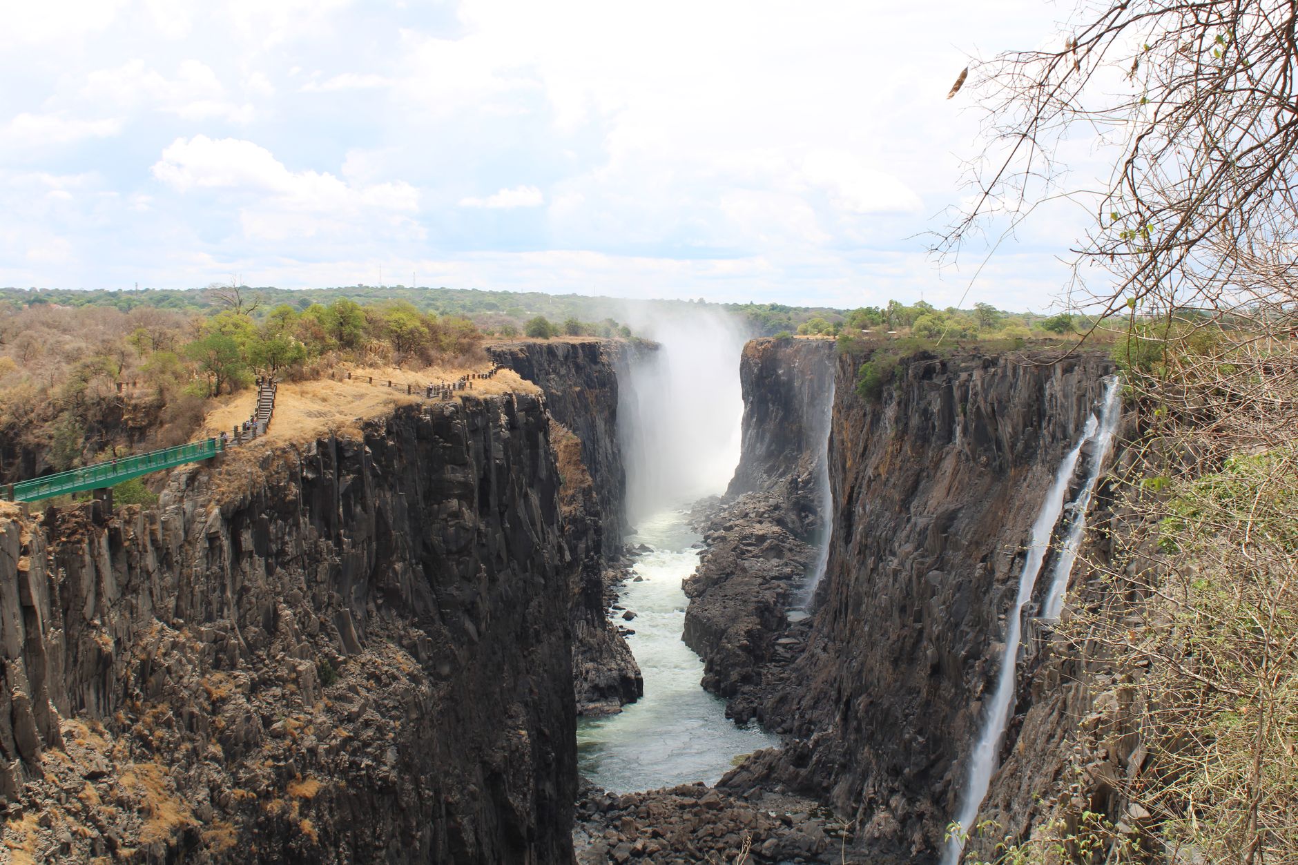 View across the Victoria Falls gorge, dark basalt cliffs largely bare with a single fall on the right and a walkway on the rim
