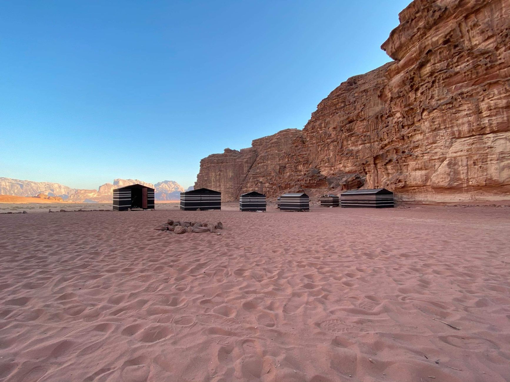 A row of black and white striped Bedouin tents on red sand below a sandstone cliff in Wadi Rum