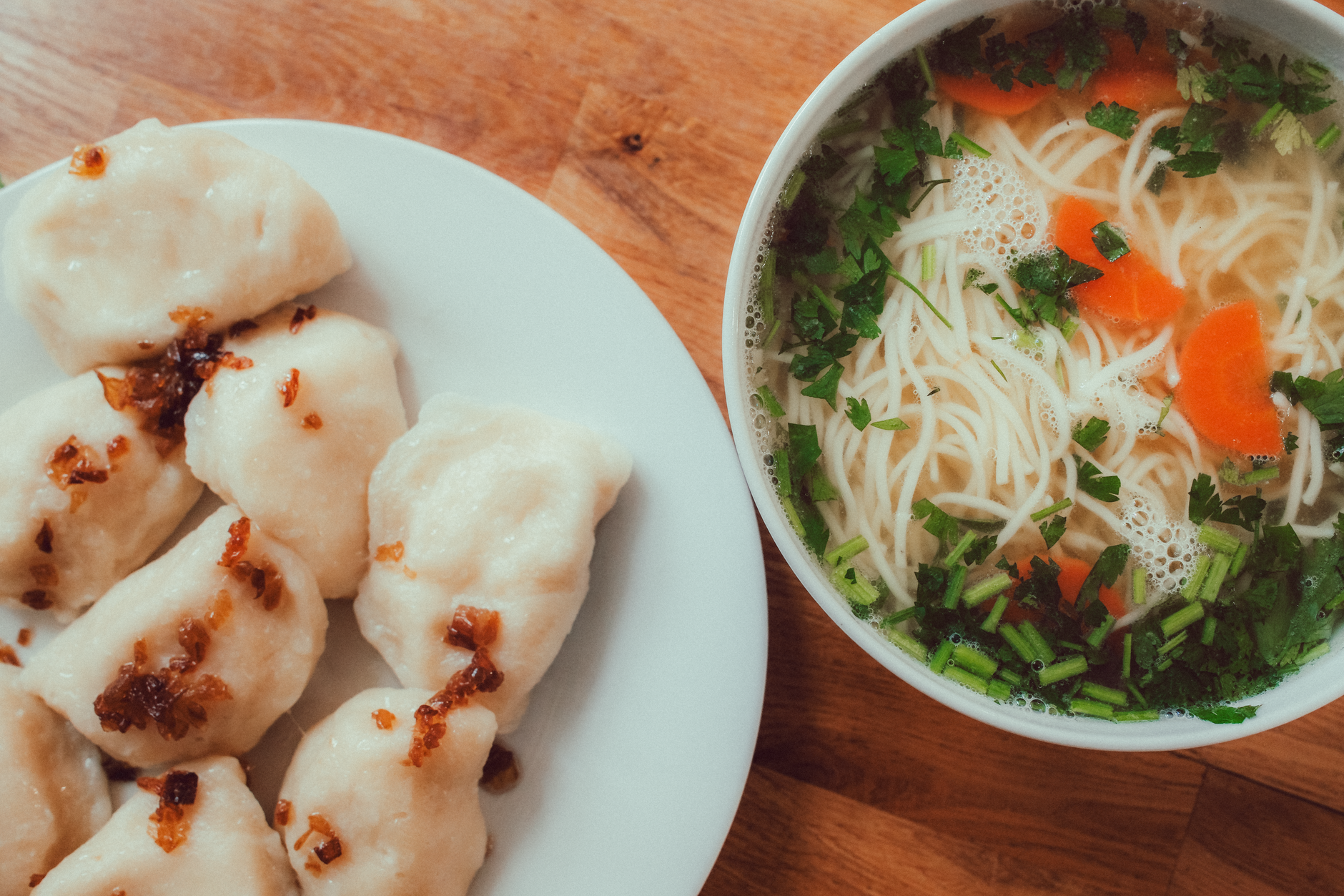 A plate of pierogi topped with fried onions beside a bowl of noodle soup