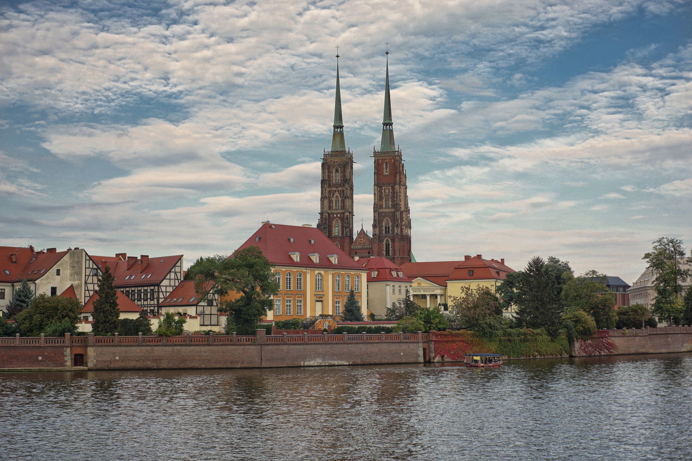 The twin spires of St John the Baptist Cathedral on Ostrów Tumski seen across the Oder, with red-roofed houses on the bank under a cloudy sky